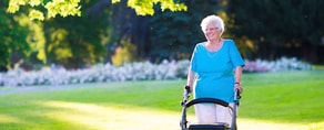 An elderly woman in a blue shirt uses a walker in a sunlit park as part of her hybrid care routine with medbridge.