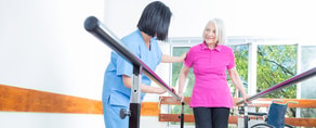 A caregiver helps an elderly woman walk between parallel bars in a well-lit room with a nearby wheelchair.