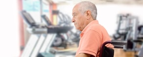 An elderly man in a wheelchair participates in a gym session, facing treadmills, as part of his medbridge routine.