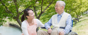 A young woman kneels by a smiling elderly man in a wheelchair, with trees and a river behind them.