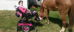 A person in a pink motorized wheelchair reaches toward a brown horse wearing a pink halter outside medbridge.