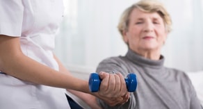 A healthcare worker assists an elderly woman with exercise using a blue dumbbell in a Medbridge session.