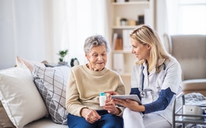 A healthcare worker explains Medbridge tablet features to an elderly woman holding a pill bottle on a sofa.