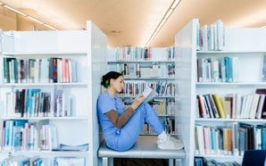 A person in blue scrubs reads a healthcare education book on a bench in the library.