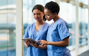 Two healthcare professionals in blue scrubs discuss medbridge software on a tablet in a bright hallway.