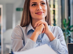 In a bright office, someone at a desk smiles with hands clasped, looking at another person.
