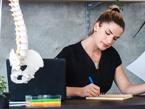 A person writing at a desk in an office decorated with anatomical posters, a spine model, and plants.
