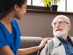 A Medbridge nurse in blue scrubs converses with an elderly man on a sofa.
