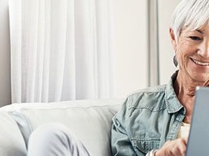 Smiling older woman with white hair uses a tablet on a softly lit couch, wearing a light shirt and earrings.