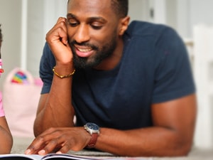 A man and a girl in a pink shirt lie on the floor smiling, reading a book together.