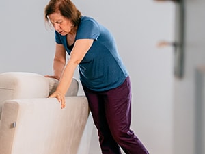 A woman in a blue shirt and dark pants holds a beige chair for support in a room, seen through a doorway.