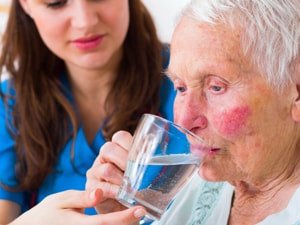 A Medbridge caregiver in a blue uniform helps an elderly person drink water.