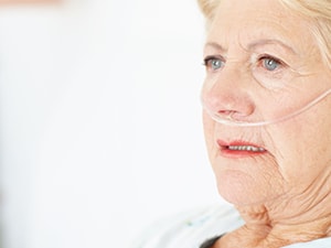 An elderly woman with short blonde hair wearing a nasal cannula looks ahead with a neutral expression in a well-lit room.
