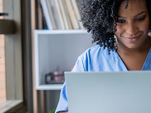 Home health clinician in scrubs reviewing OASIS documentation on a laptop, representing OASIS accuracy and training.