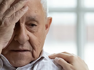 A distressed elderly man sits with his hand on his forehead, comforted by someone next to him touching his shoulder.