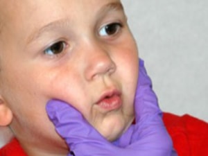 Child in a red shirt with cheeks gently held by purple-gloved hands.
