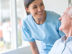 A Medbridge nurse smiles at a senior man in a wheelchair near a sunny window.