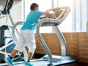 A child in blue and white stands on a treadmill in a bright gym with various exercise machines.
