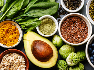 An array of fruits, vegetables, nuts, and seeds on a table with grapefruit, apple, spinach, broccoli, avocado, blueberries, almonds, and pomegranate.