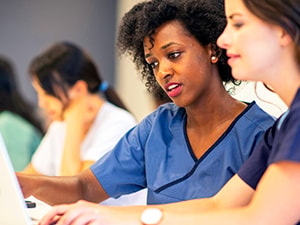 Two women in medical scrubs focus on a laptop screen, with others working in a medbridge classroom.