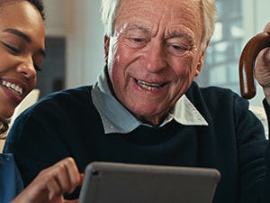 A young woman shares a tablet with an elderly man holding a cane, both smiling in a living room.