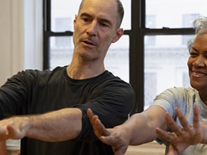 Two people exercise indoors with arms extended, behind them are a window and exercise balls.