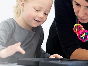A child and an adult with a colorful necklace focus on a black circular object on a table during Medbridge's activity.