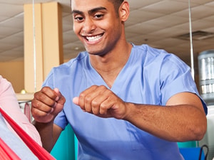 An elderly person exercises with a red resistance band, assisted by a smiling healthcare worker in medbridge scrubs.