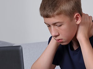 Teen boy in a navy shirt looks at a laptop with his hands on his neck.