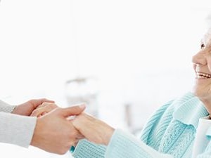 An elderly person in a light blue sweater smiles while holding hands with someone in white in a bright, blurred background for medbridge.