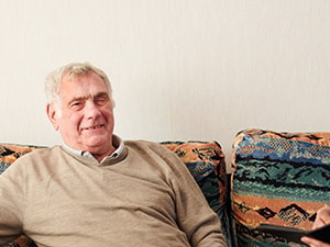 A young man in a blue uniform with a clipboard talks to an elderly man on a patterned sofa.