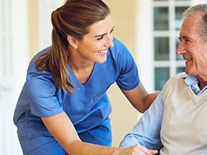 Smiling clinician in blue scrubs speaking with an older adult man in a wheelchair outside a care facility.