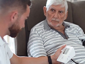 Home health clinician uses a blood pressure monitor while discussing heart failure care with an older male patient.
