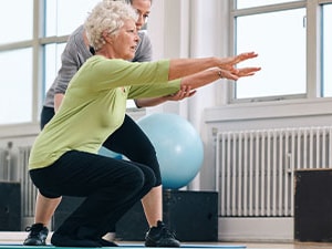 Older adult performing a balance squat with support from a physical therapist in a gym setting for fall prevention training.