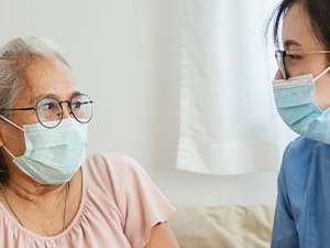 An elderly woman and a nurse in a blue uniform, both masked, talk indoors.