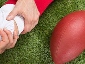 Person supporting athlete's leg in white pants and red socks near a football on grass.