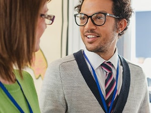 Two professionals with name badges converse near a wall of maps, both wearing glasses.