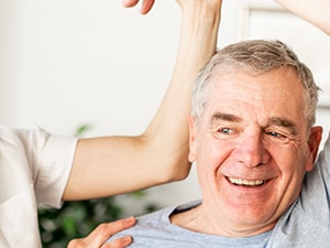 A Medbridge therapist helps an elderly man with arm exercises in a bright room.