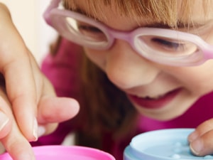A child with glasses happily plays with containers, helped by an adult hand.