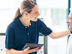 Female clinician smiling and holding a tablet while talking to an older male patient during a check-in, representing patient engagement.