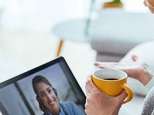 Woman sits on a couch holding a coffee mug while speaking to a healthcare provider via telehealth video call on a laptop.