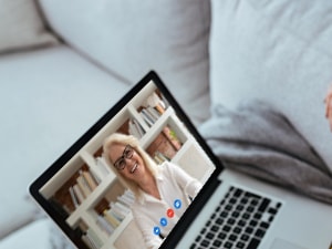 A woman in a yellow sweater is on a video call via laptop with another person smiling in front of bookshelves, using Medbridge.