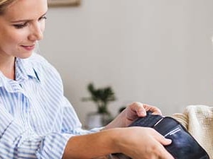 In a bright living room, a caregiver helps an elderly woman use a blood pressure monitor.