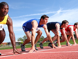 Five male athletes in numbered jerseys are in starting position on a sunny outdoor track.