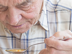 Elderly man in plaid shirt eating soup indoors by a window.