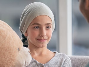A young child with a headscarf holds a teddy bear, sitting opposite a woman in a medbridge uniform.