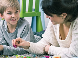 A woman and child build with blocks on a rug, with colorful chairs behind them.