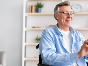 A smiling older man in a wheelchair uses a smartphone, with books and plants in the background.
