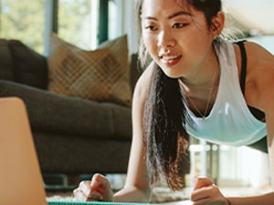 A woman does a plank exercise indoors while watching a medbridge video on a laptop.