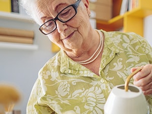 Elderly woman with glasses and floral shirt watering indoor plants.